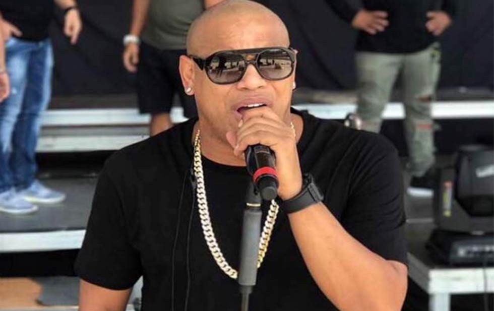 Hombre cantando en micrófono, con gafas y collar.