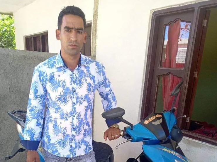 Hombre junto a moto azul con camisa estampada.