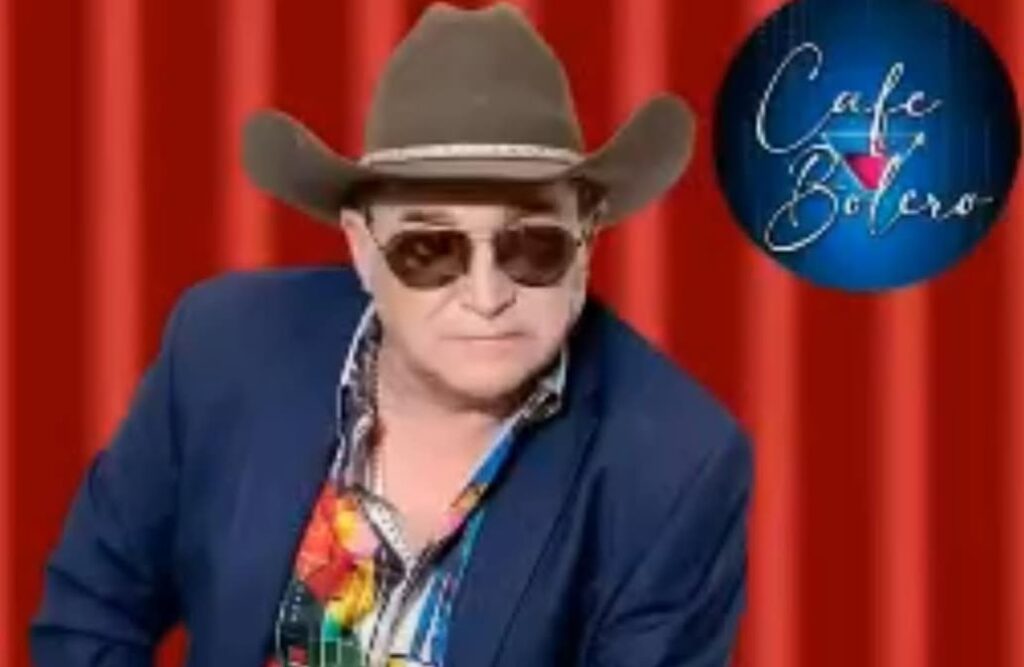 Hombre con sombrero, gafas y chaqueta frente a cortina roja