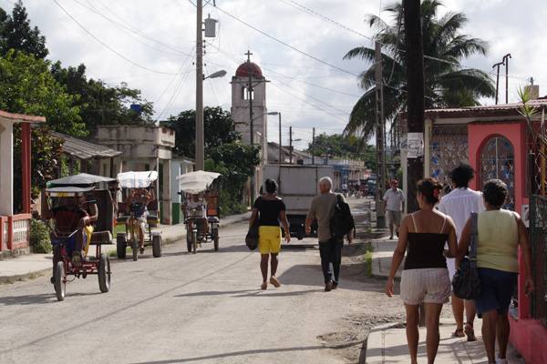 Calle con peatones y bicitaxis, iglesia al fondo.