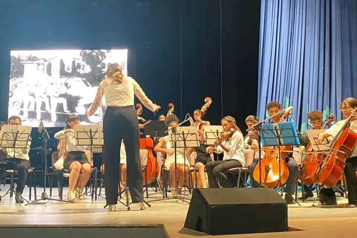 Orquesta tocando en concierto con directora de espaldas.