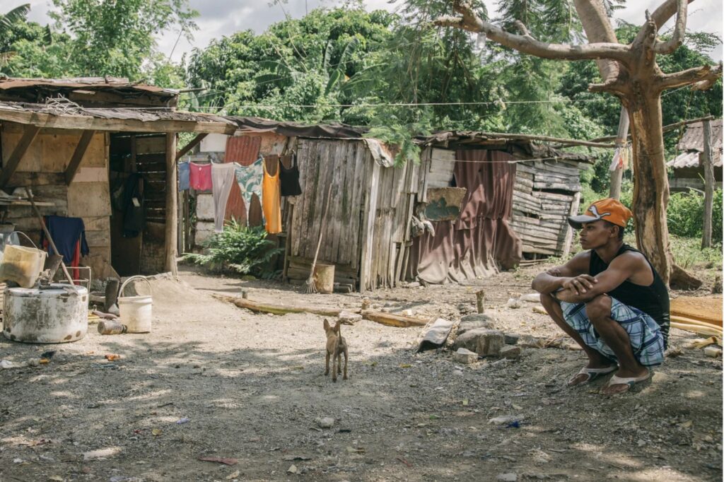 Hombre sentado frente a chabola con perro cercano.