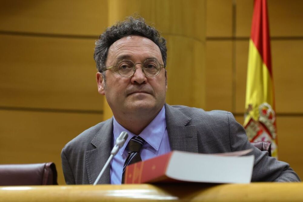 Hombre con traje en oficina gubernamental, bandera española detrás.