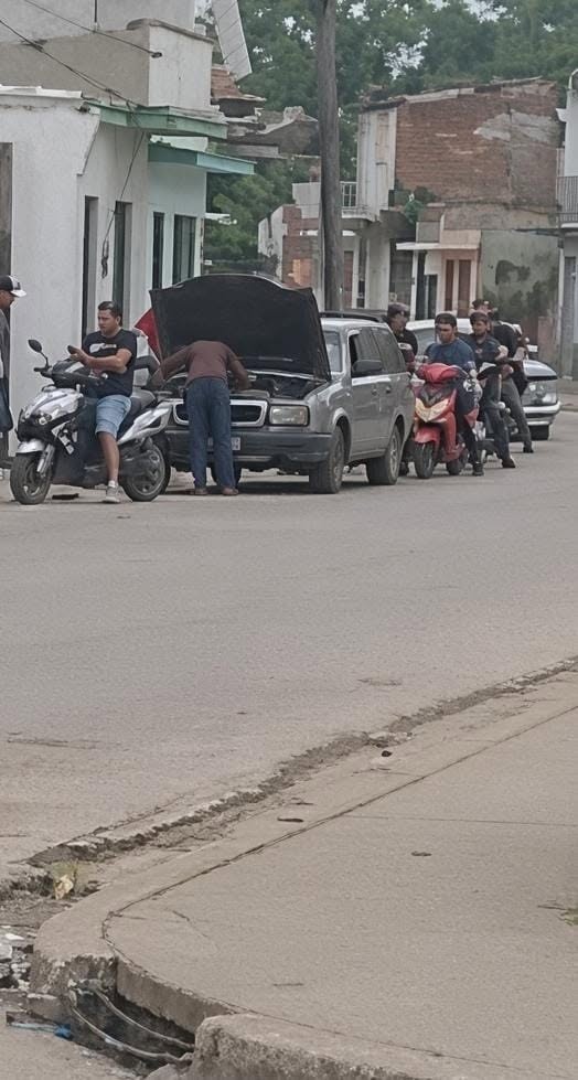 Hombre reparando coche plateado en una calle concurrida.