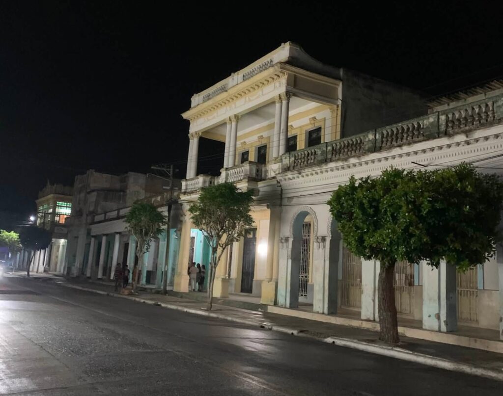 Calle nocturna con edificios coloniales iluminados y árboles pequeños.