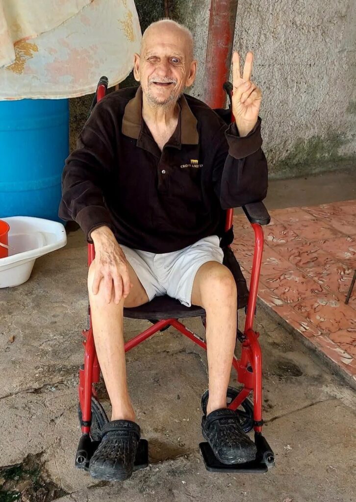 Anciano en silla de ruedas sonriendo con signo de paz.