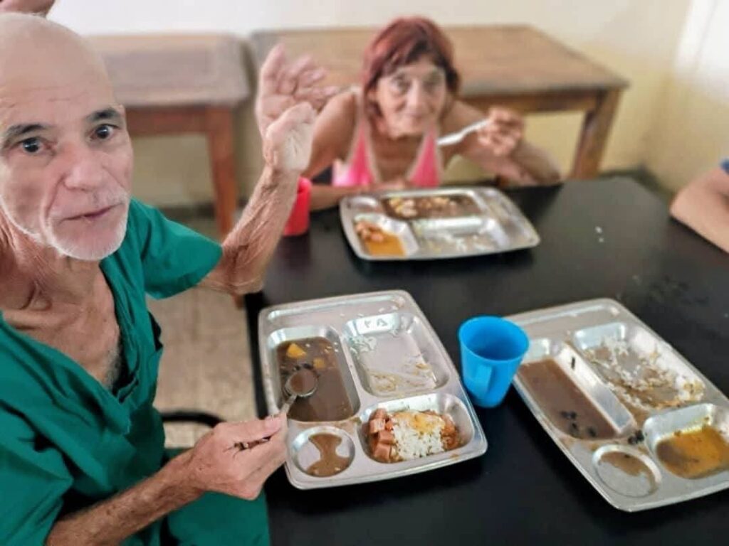 Dos personas mayores comiendo de bandejas metálicas en un comedor.