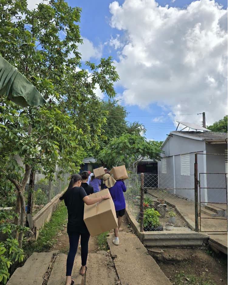 Personas cargando grandes cajas de cartón por un camino rural.