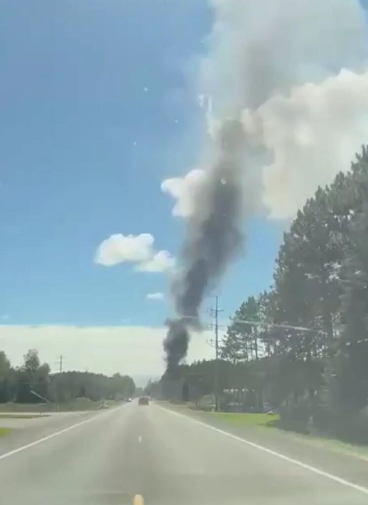 Columna de humo negro surgiendo tras árboles junto a carretera.