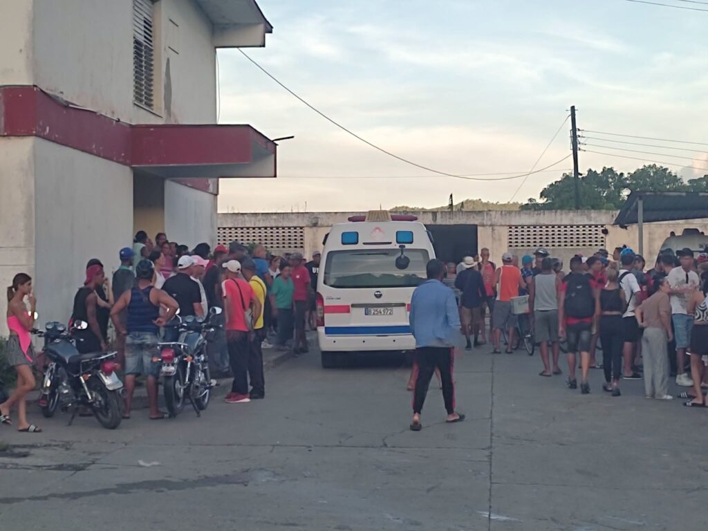 Multitud reunida ante un edificio junto a una ambulancia blanca.