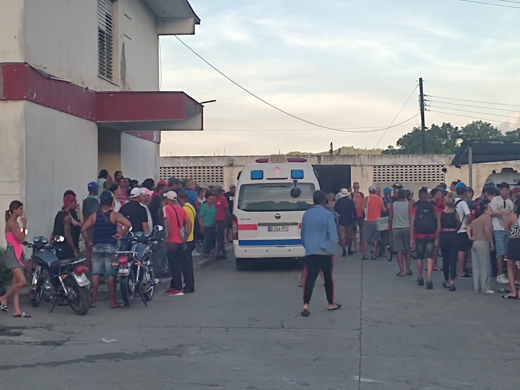 Multitud reunida ante un edificio junto a una ambulancia blanca.