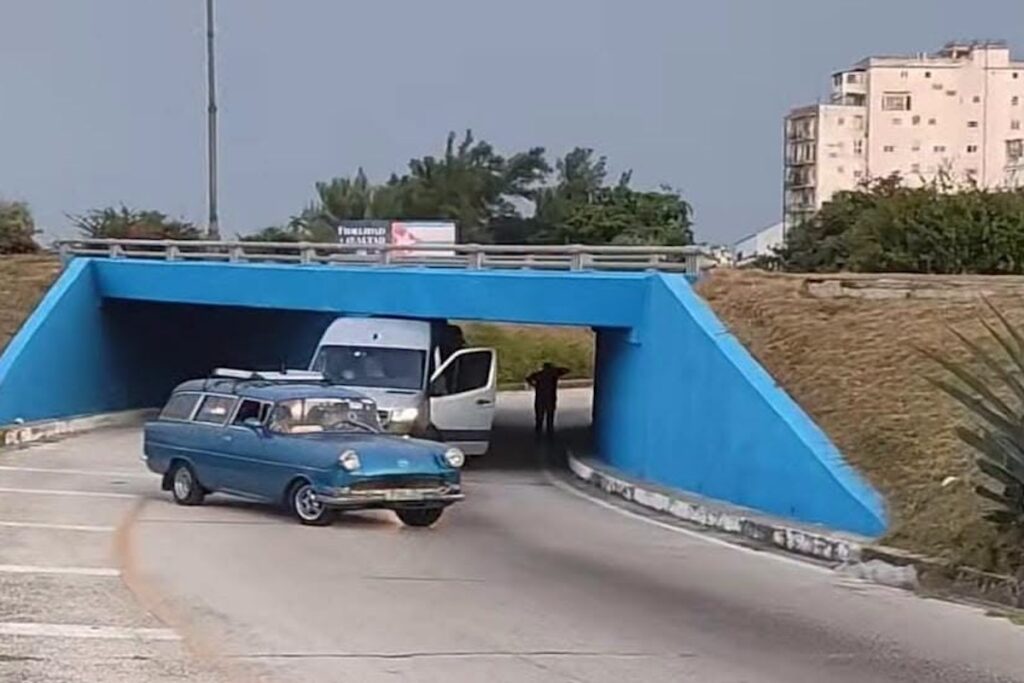 Furgoneta blanca atascada bajo un puente azul bajo.