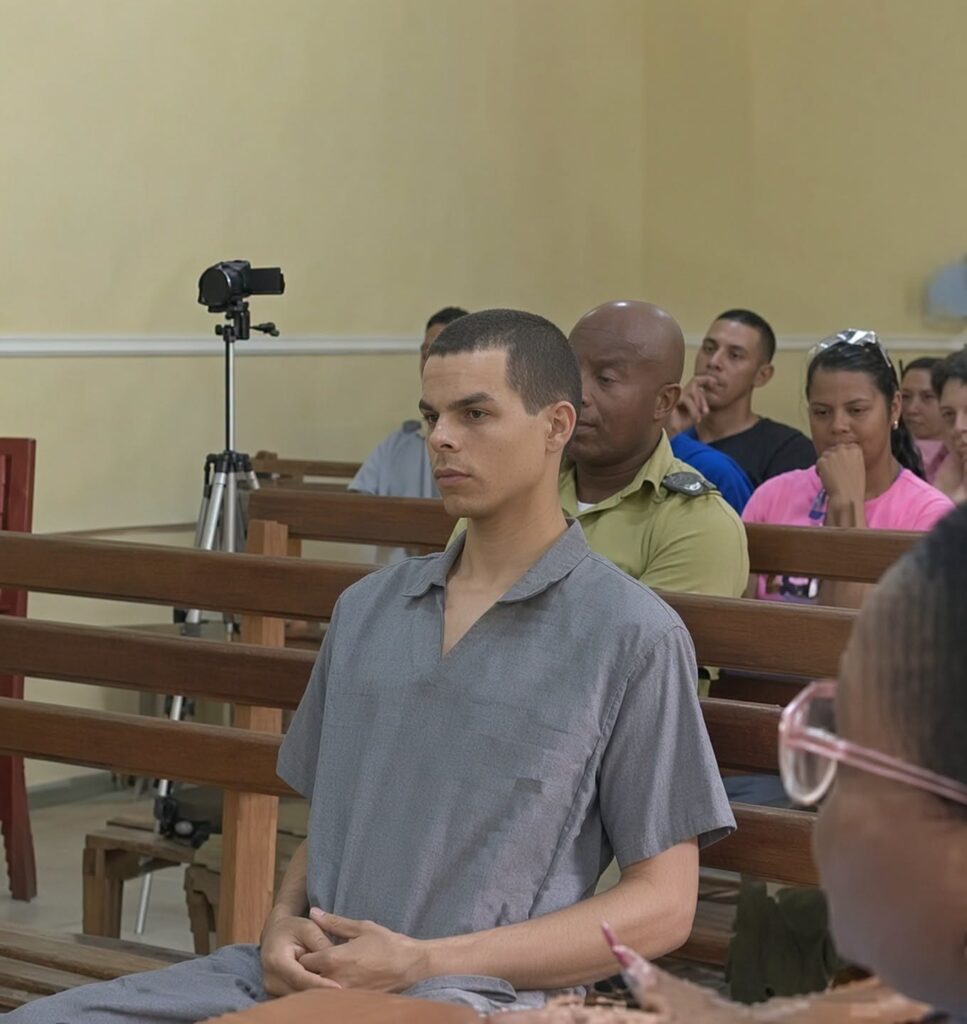 Hombre joven con camisa gris sentado en una sala judicial.