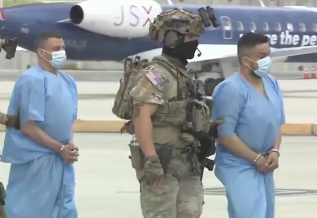 Soldado escoltando a dos detenidos esposados junto a un avión.