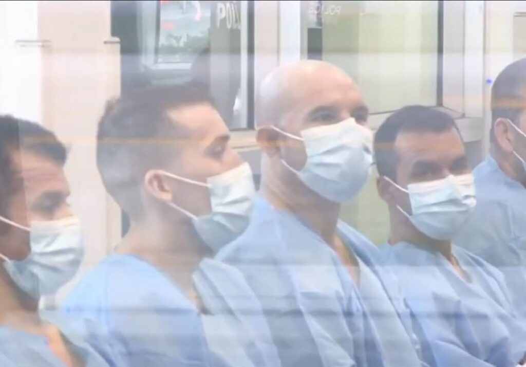 Hombres con batas azules y mascarillas blancas tras un cristal.