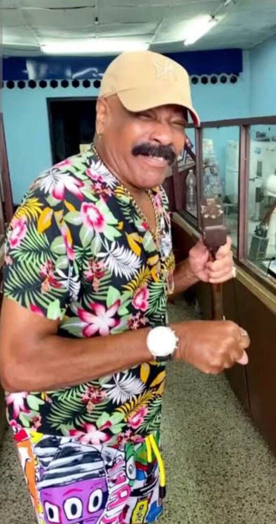 Hombre sonriente con bigote, gorra beige y camisa floral.