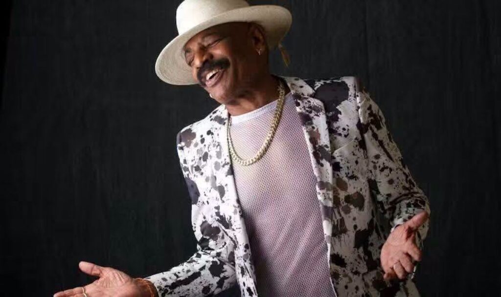Hombre con sombrero y chaqueta estampada sonriendo alegremente.