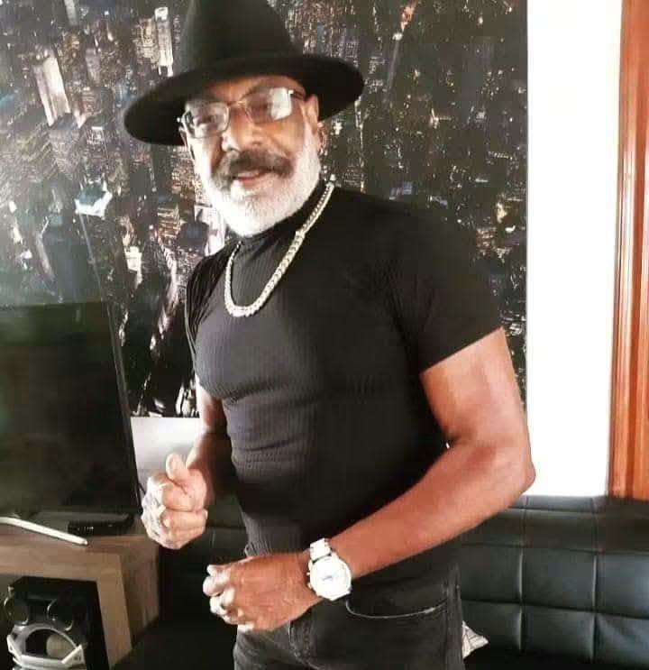 Hombre mayor con barba gris, sombrero negro y cadena plateada.