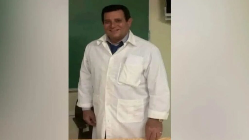 Hombre sonriente con bata blanca frente a una pizarra verde.