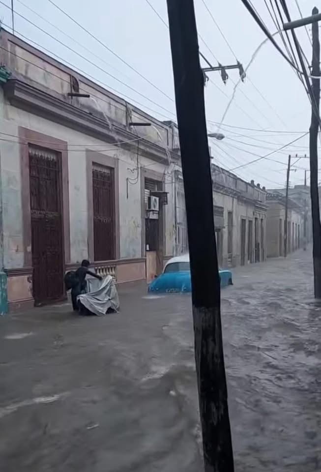 Calle inundada con coche clásico azul y persona empujando carrito.