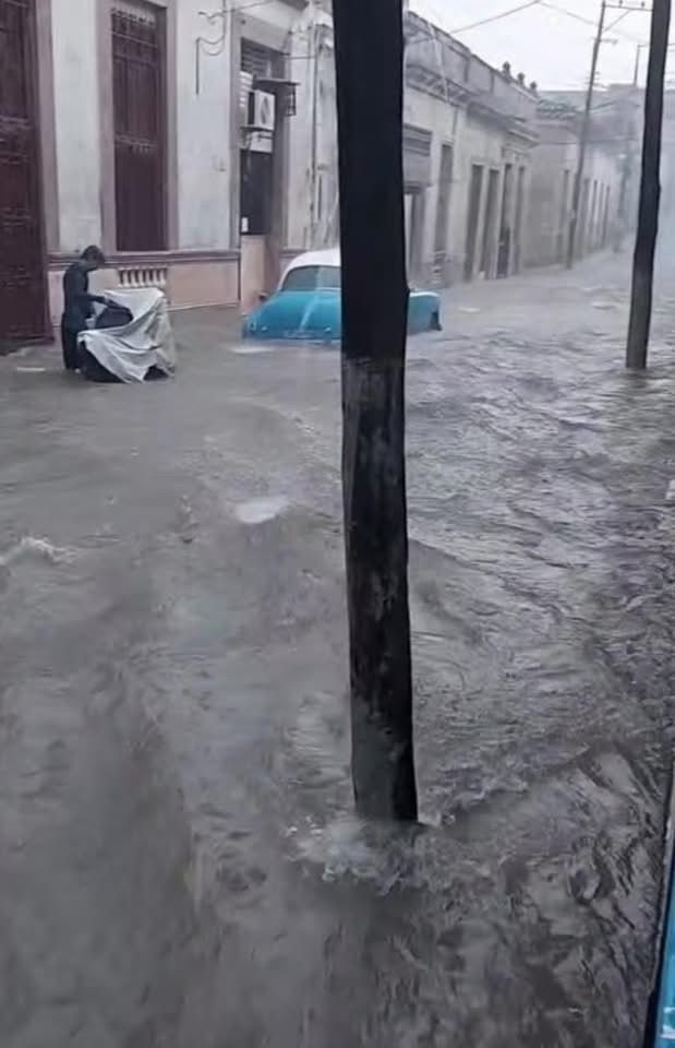 Calle inundada con un coche clásico azul sumergido parcialmente.